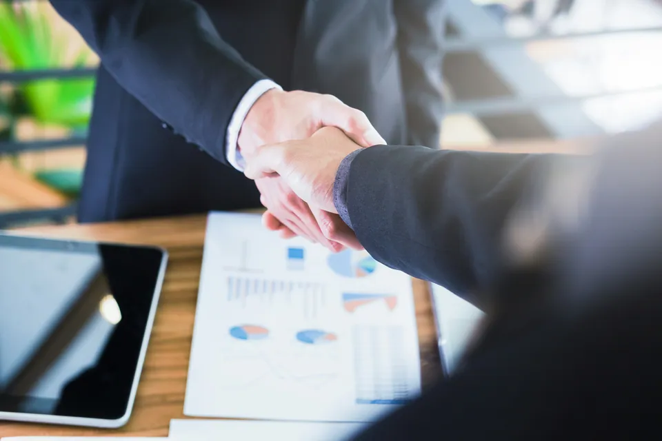 Two businesspeople shaking hands over a desk with printed charts and a tablet