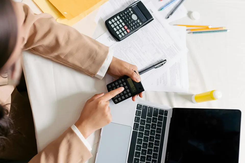 Overhead view of hands using a calculator next to a laptop and paperwork