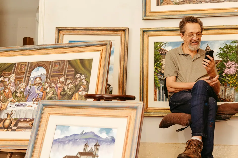 Smiling art dealer seated among framed paintings while looking at his phone