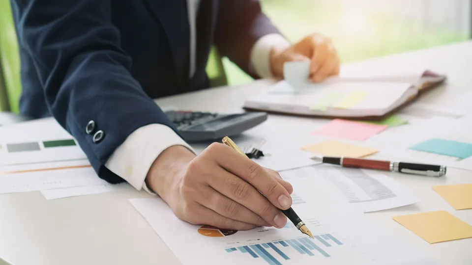 Hand with a pen reviewing printed financial charts and sticky notes