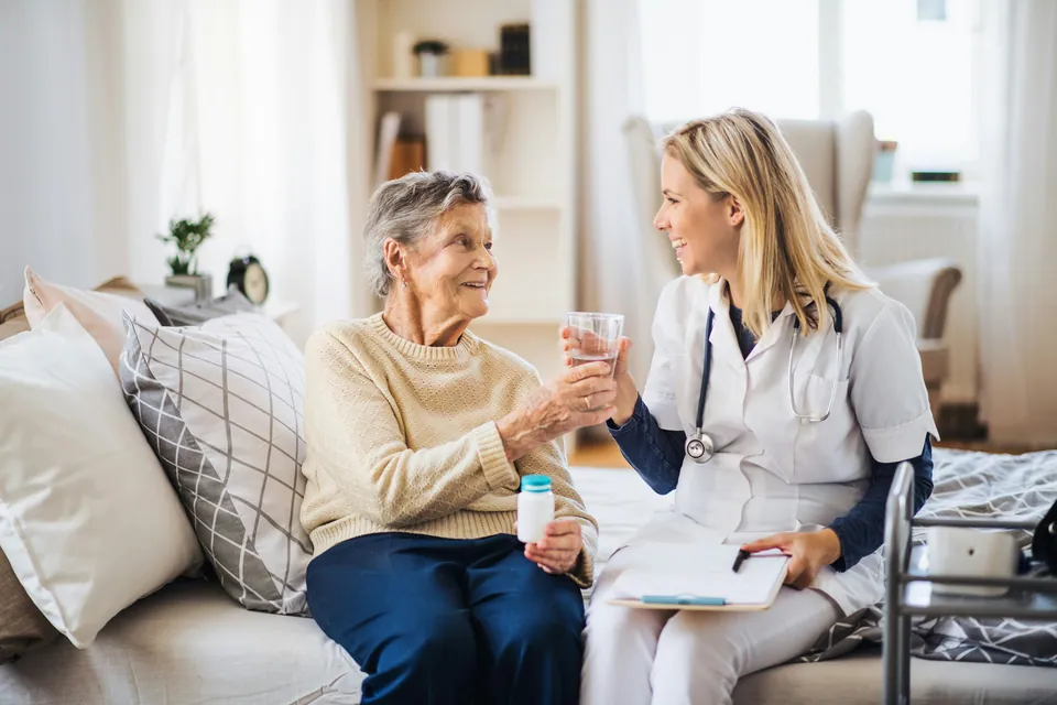 Nurse talking with an older woman holding a glass of water on a sofa