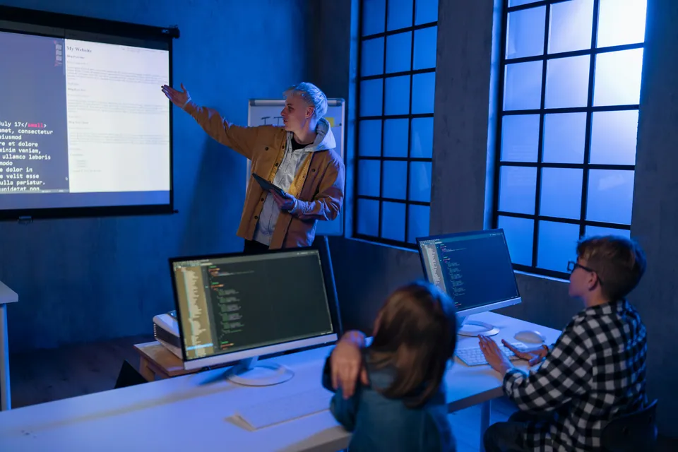 Instructor pointing at code on a projector screen while students work at monitors