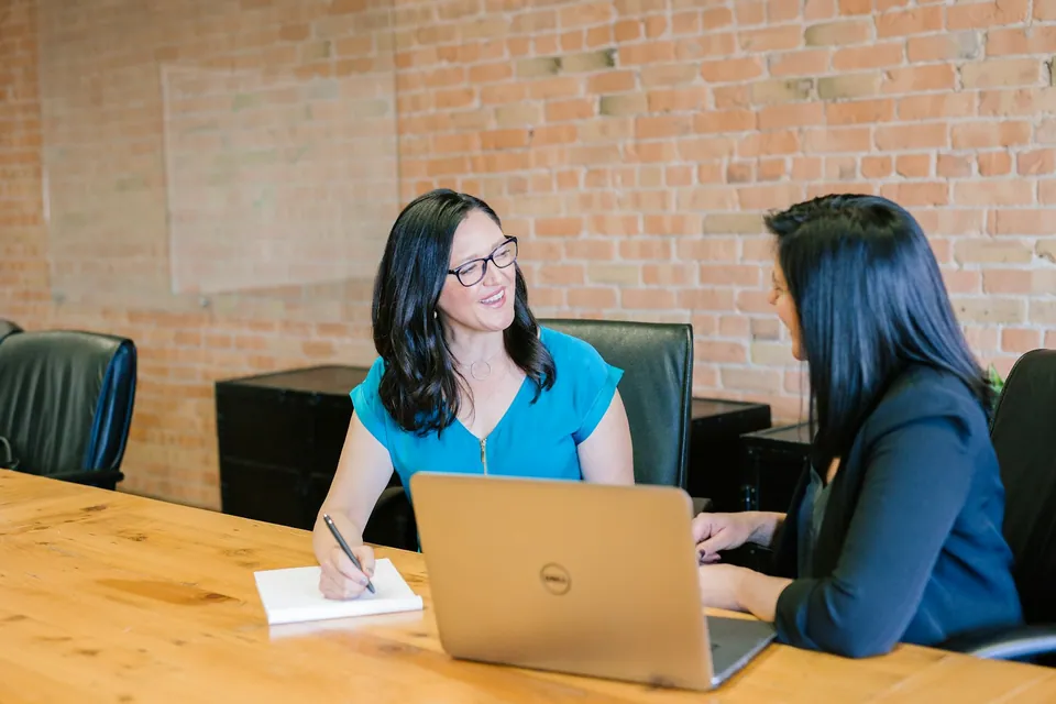 Two women talking at a wooden table with a laptop and notepad