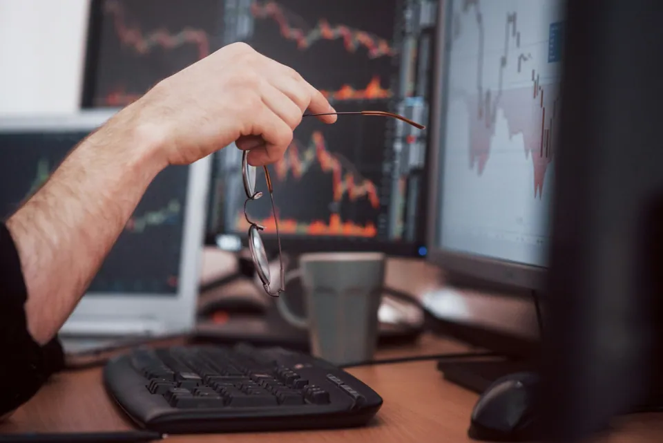 Trader holding glasses in front of multiple screens of market charts