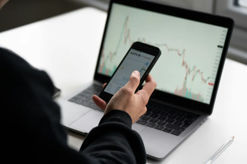 Person checking a stock chart on a phone in front of a laptop
