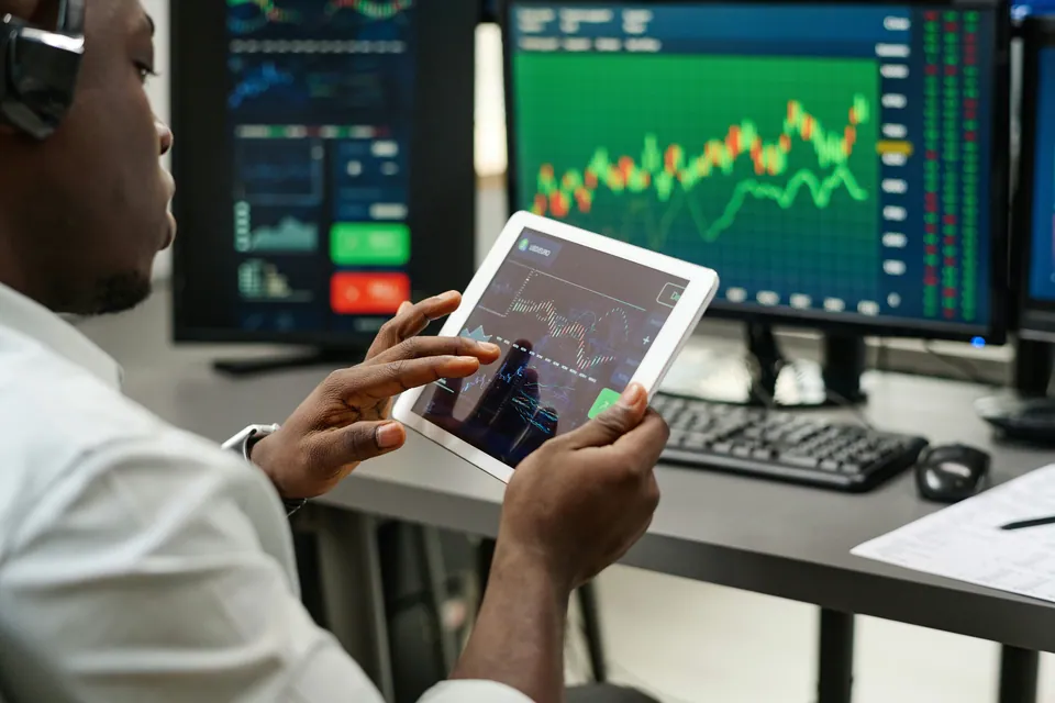 Trader holding a tablet with a stock chart in front of trading screens