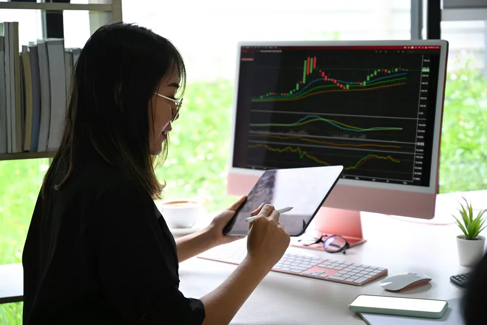 Woman reviewing a stock chart on a tablet in front of a desktop monitor
