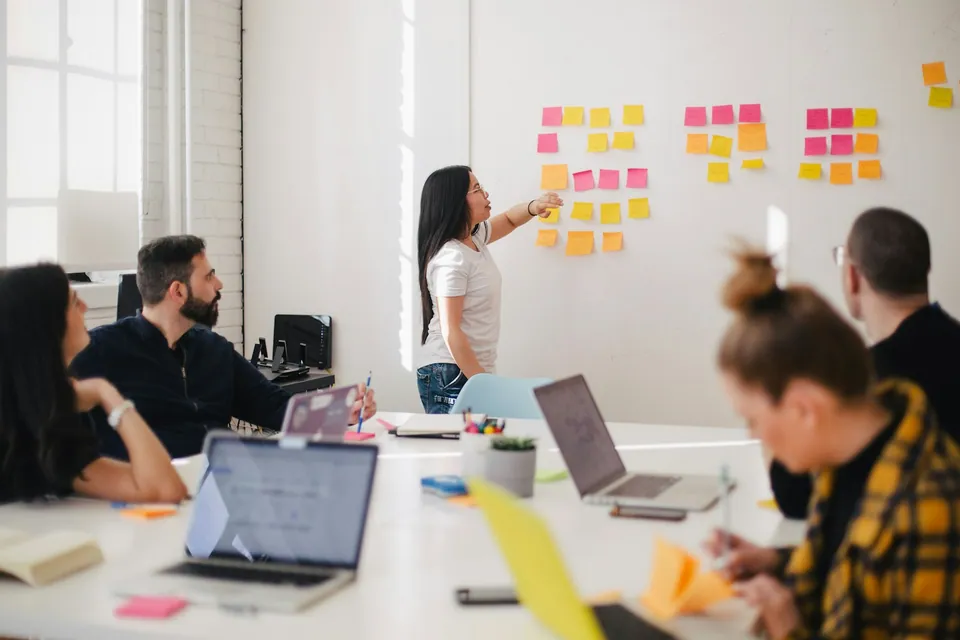 Woman presenting sticky notes on a wall to a team with laptops