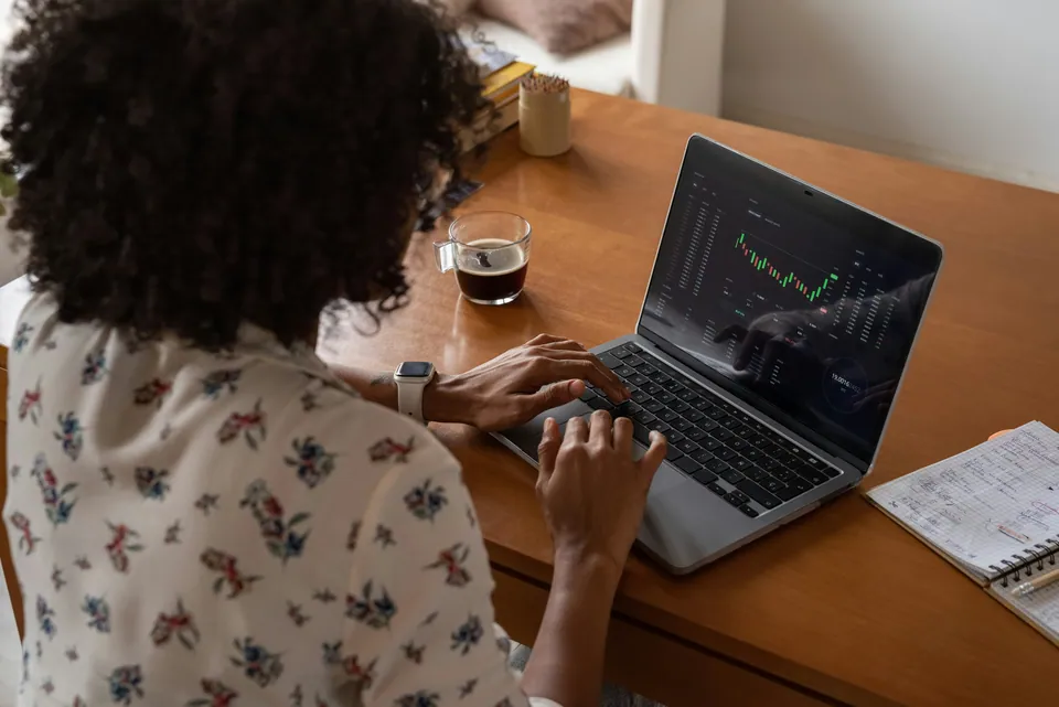 Woman typing on a laptop showing a trading chart at a wooden desk