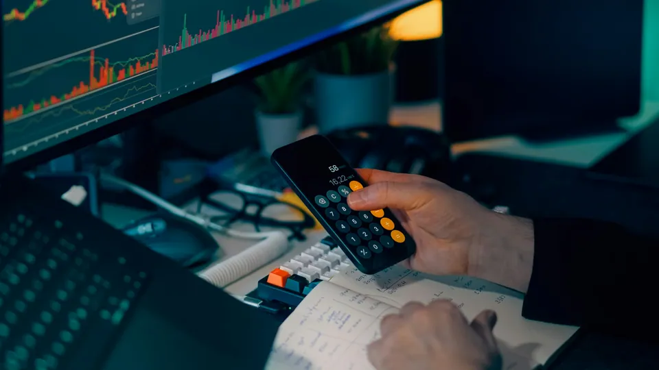 Hands using a phone calculator in front of trading screens