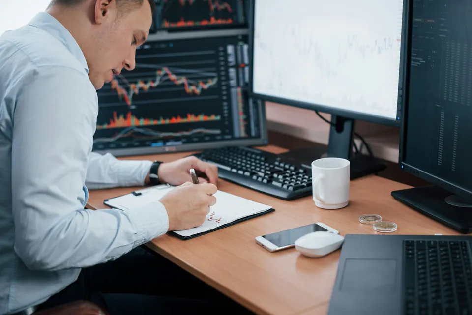 Trader taking notes in front of multiple chart monitors