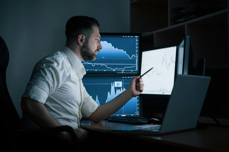 Trader pointing at chart monitors in a dark office