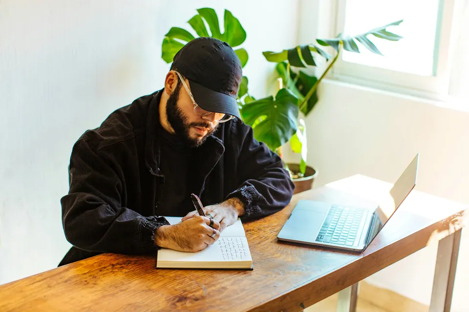 Man in a cap writing in a notebook beside a laptop and plants