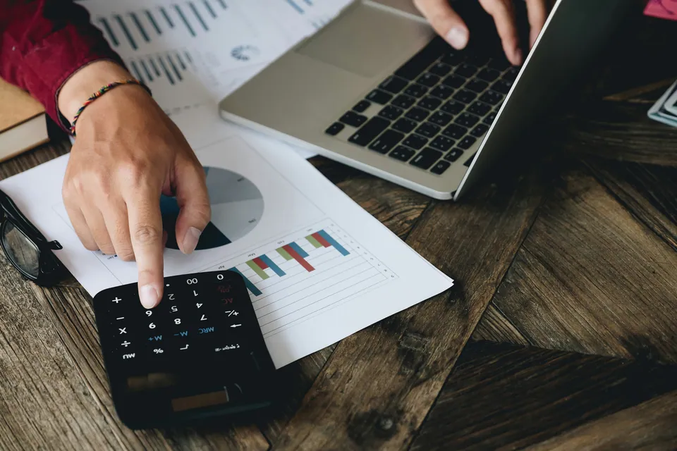 Hand using a calculator over printed charts beside a laptop