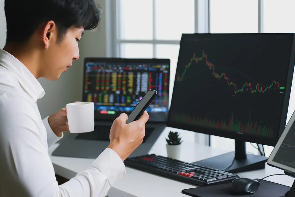 Young trader checking his phone in front of stock chart monitors