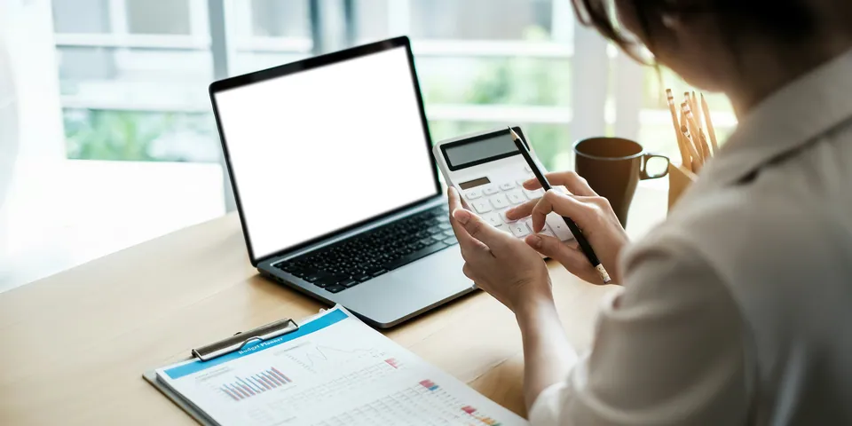 Woman using a calculator beside a laptop and printed paperwork