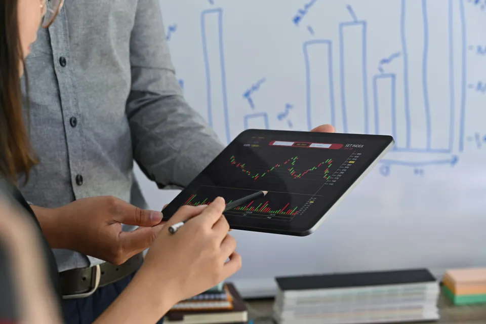 Two people reviewing a stock chart on a tablet by a whiteboard
