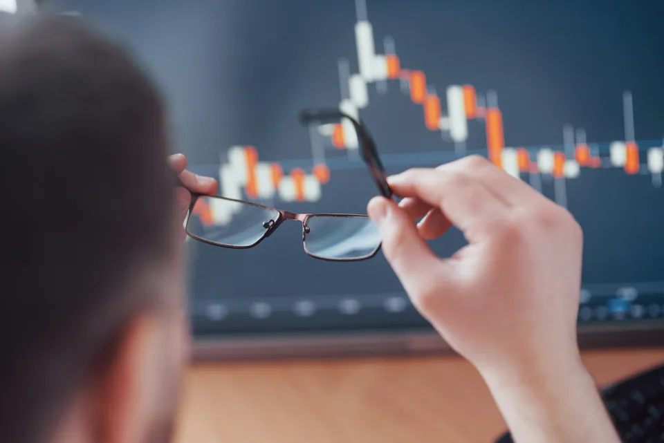 Person holding glasses in front of a blurred candlestick chart