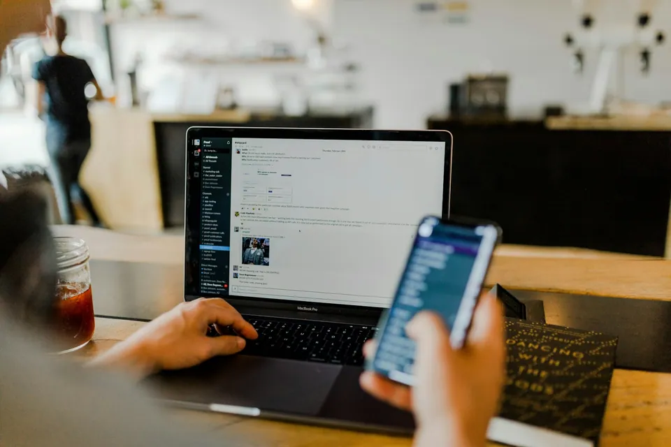 Person holding a phone in front of a laptop at a cafe table