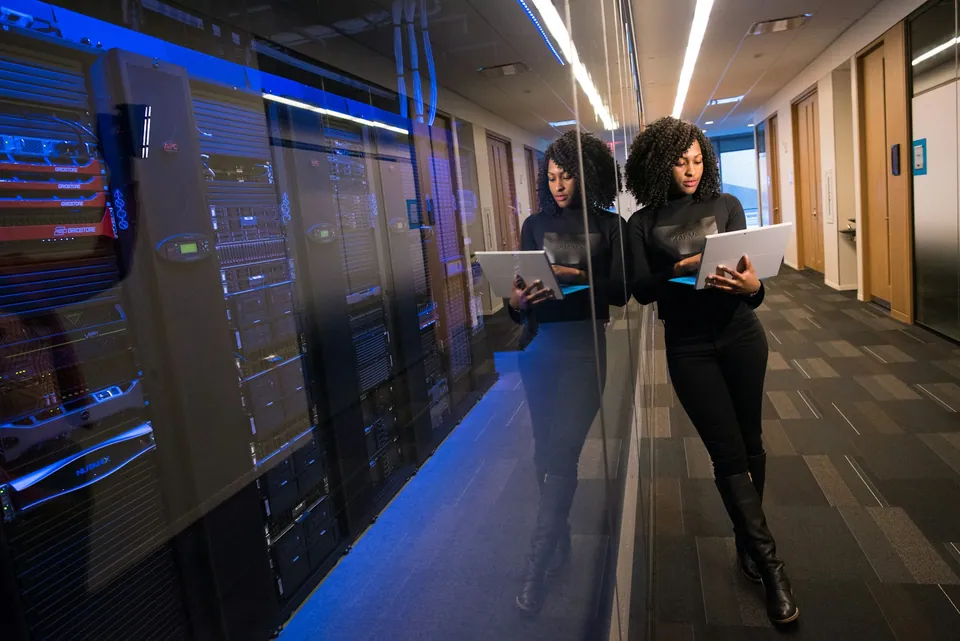Two women with laptops walking through a server room aisle