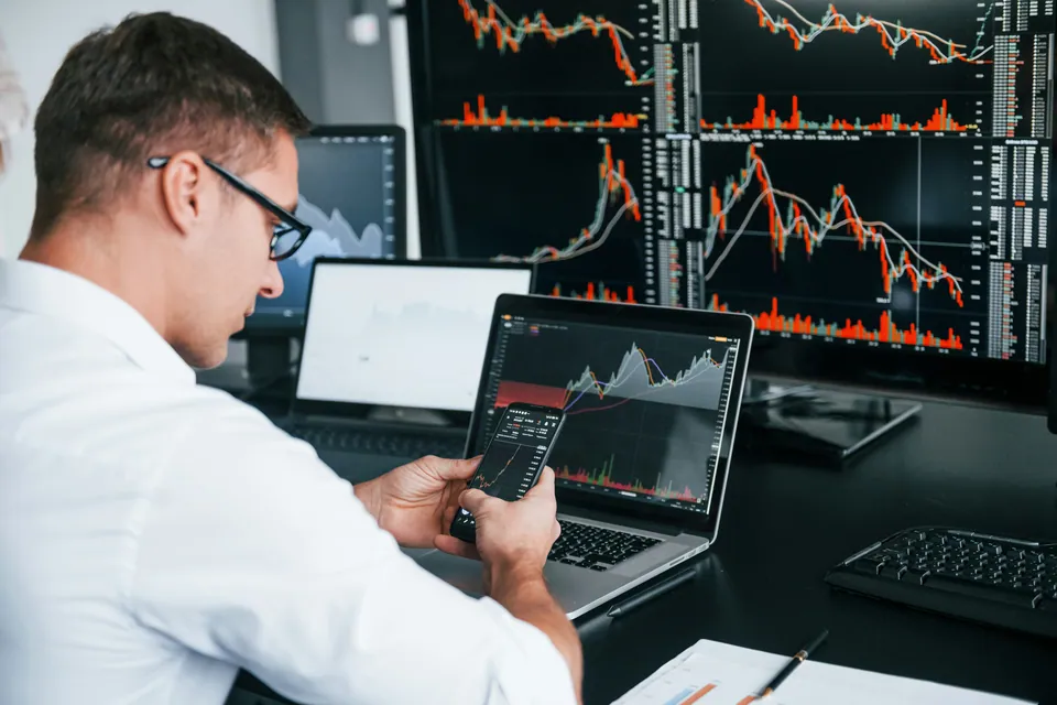 Trader using a phone calculator in front of stock chart screens