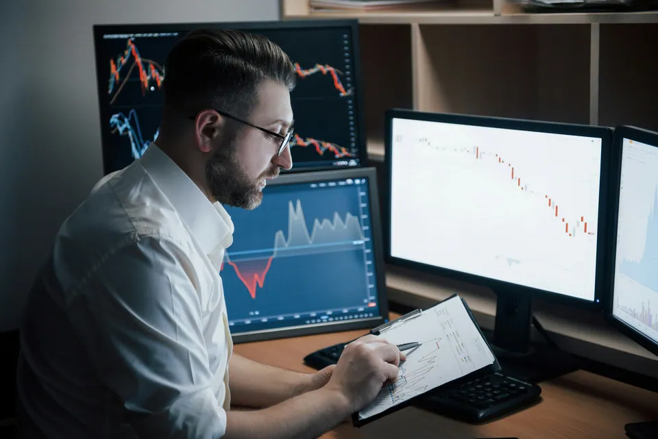 Trader taking notes in front of multiple chart monitors