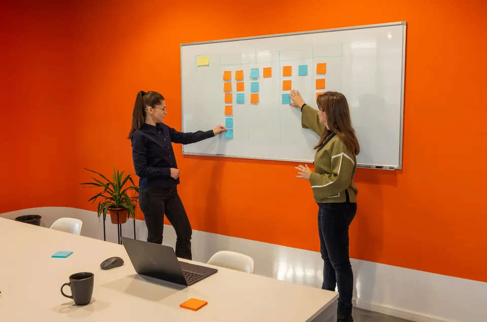 Two women arranging sticky notes on a whiteboard against an orange wall