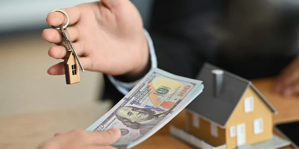 Hand passing house keys and cash next to a model house