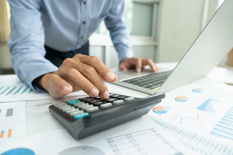 Hand using a calculator over printed charts beside a laptop
