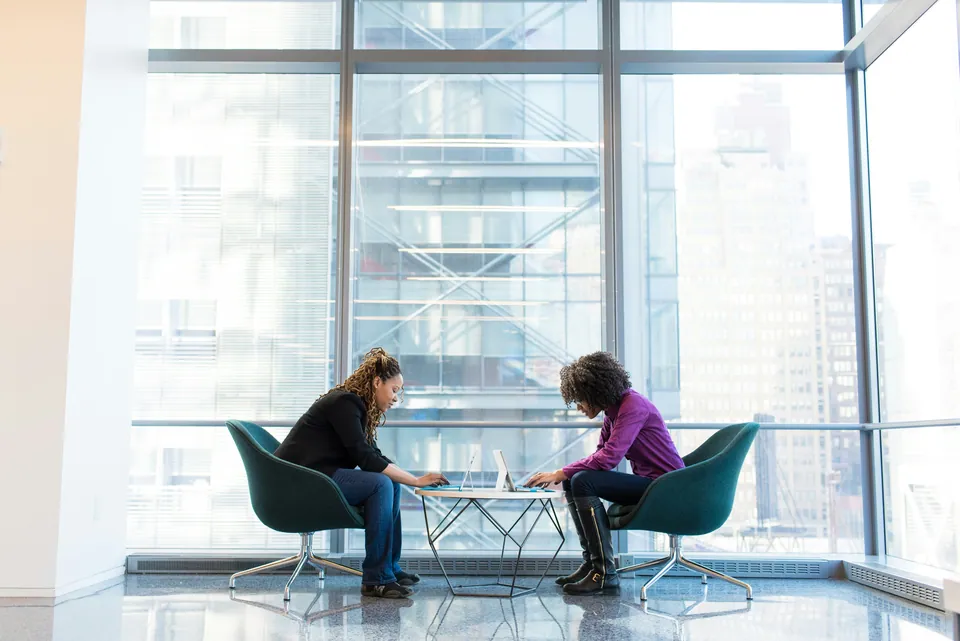 Two women talking at a small table by tall office windows