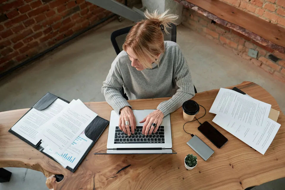Woman typing on a laptop at a wooden desk with printed reports