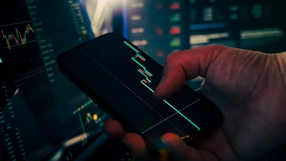 Hand holding a phone showing a price chart in front of trading screens