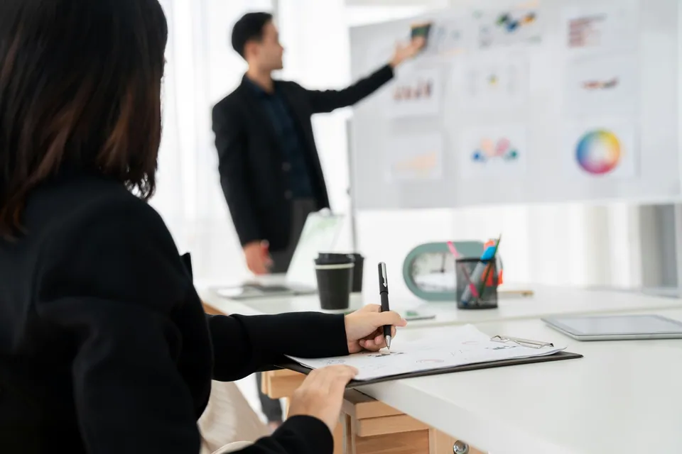 Presenter pointing at charts on a whiteboard while a colleague takes notes