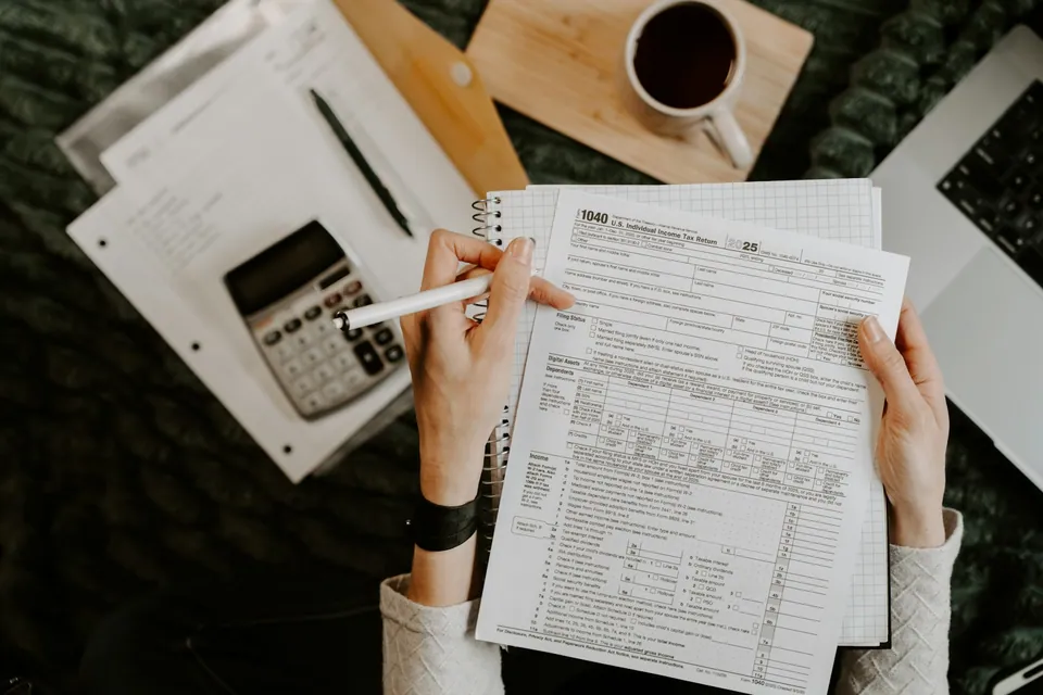 Person filling out a tax form beside a calculator, coffee and laptop