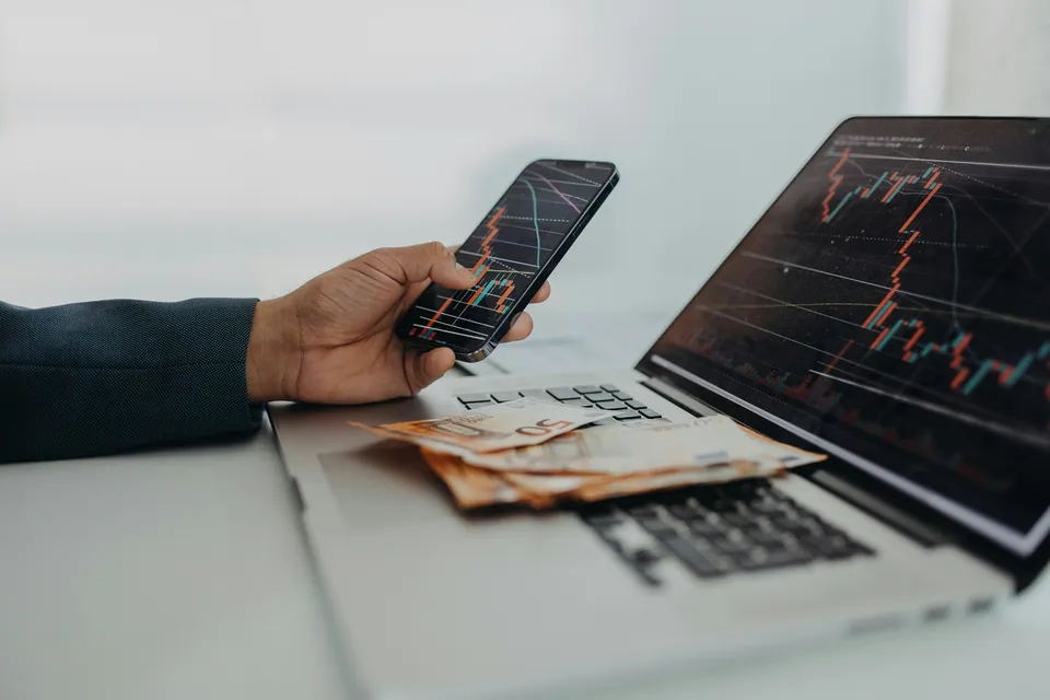 Hand holding a phone with a price chart beside a laptop and cash