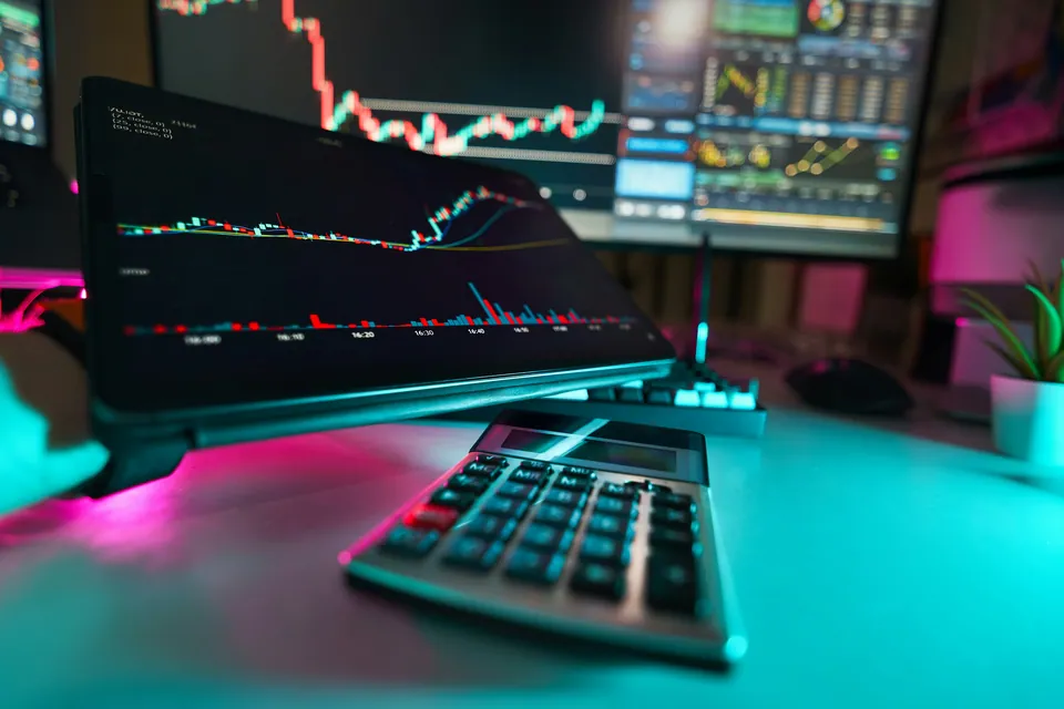 Calculator in front of laptops showing trading charts under neon light