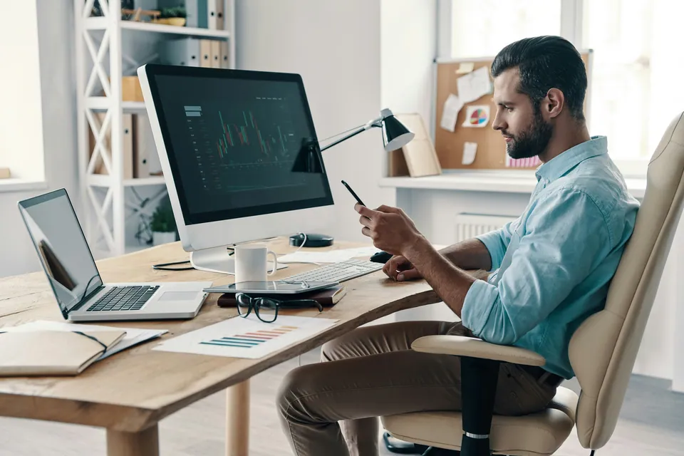 Man checking his phone at a home office desk with a monitor and laptop