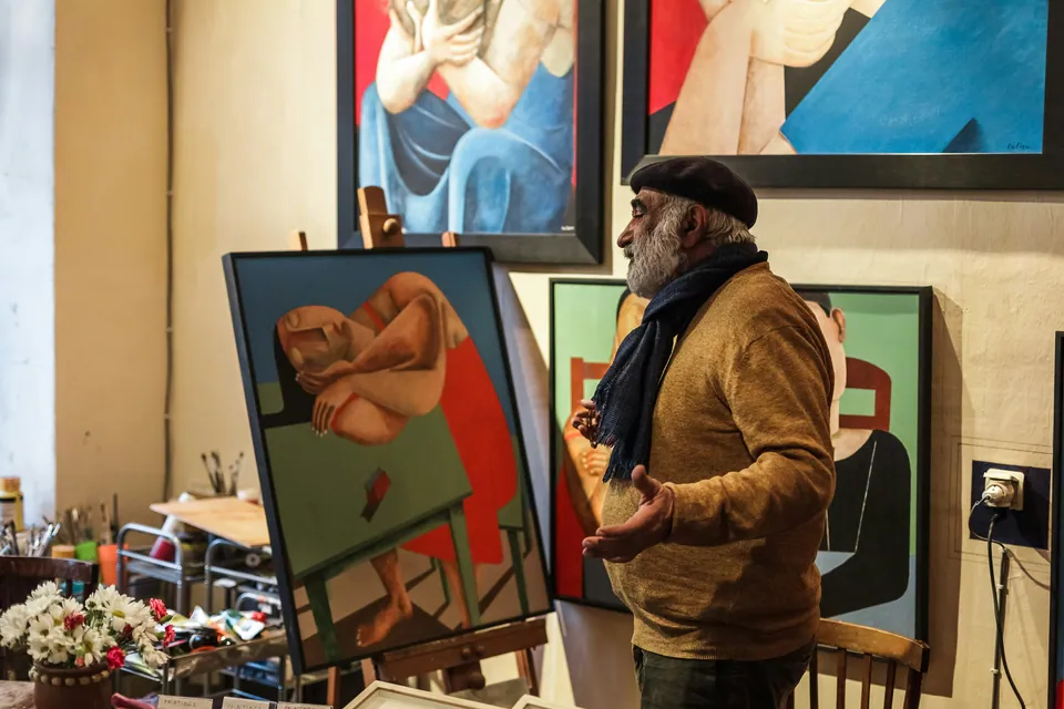 Older painter talking in a studio full of colorful canvases