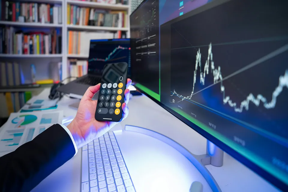 Hand holding a phone calculator in front of a monitor with price charts