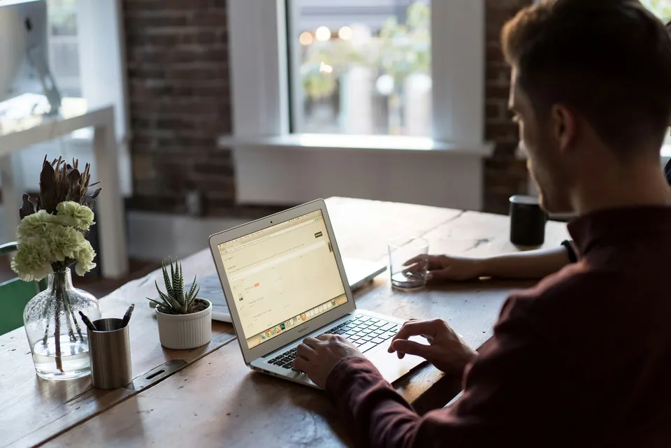 Man typing on a laptop at a wooden table by a window