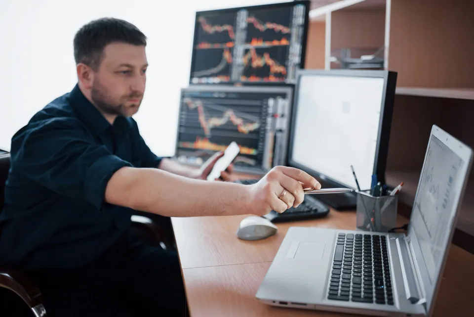 Trader pointing at a laptop in front of chart monitors