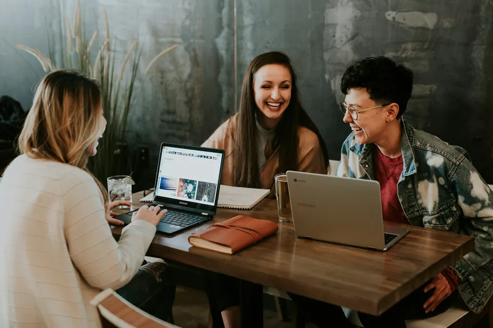 Three friends laughing around a cafe table with laptops