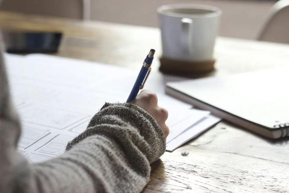 Hand writing notes beside a coffee cup at a wooden desk